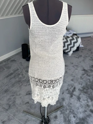 Vit virkad strandklänning - Säljer en vit virkad strandklänning med ärmlös design och rund hals. Klänningen har ett vackert spetsmönster nertill med blommiga detaljer som ger en bohemisk känsla. Perfekt att dra över bikinin på stranden eller vid poolen.