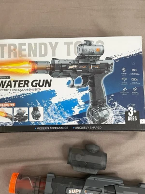 Elektrisk vattenpistol med tillbehör - Säljer en elektrisk vattenpistol i svart och grå plast med orange detaljer. Medföljer avtagbar sikte, vattenbehållare och USB-laddkabel. Pistolen har modern design och passar för vattenlekar utomhus. För barn från 3 år och uppåt.