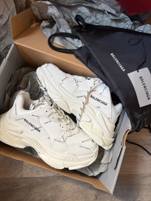 Vita Balenciaga Triple S sneakers - Chunky vita Balenciaga Triple S sneakers med svart logotyptryck över hela skon. Tjock sula, rund tå och snörning med svartvita skosnören. Kommer med originalpåse och box. Tillverkade i en mix av läder och mesh för en modern look.