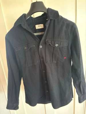 Svart overshirt från R.D.D. - Stilren svart overshirt från R.D.D. i mjukt tyg. Skjortan har två bröstfickor med lock och knappar, klassisk krage och knäppning framtill. Perfekt lager-på-lager-plagg för en avslappnad look.