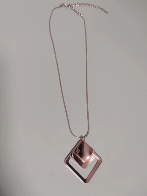 Silverfärgat halsband med geometriskt hängsmycke/berlock - Snyggt silvrigt halsband  med en geometriskt berlock bestående av tre kvadrater. Kedjan är tunn och har justerbar längd, 38 + 8 cm lång. Har bara använts ett par gånger är i fint skick (en del tunna repor). Känner inte till vad det är för typ av metall. Kan skickas med ett frimärke (22 kr).