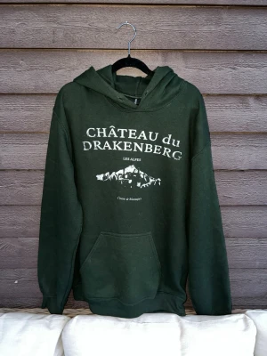 Mörkgrön hoodie Drakenberg - Mörkgrön hoodie med vit text och tryck framtill: 'CHÂTEAU du DRAKENBERG LES ALPES'. Har huva och magficka. Storlek S och gröna snören tillkommer! Hör av dig vid funderingar!