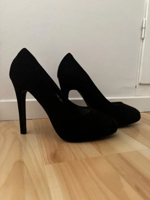 Svarta klassiska pumps med hög klack - Stilrena svarta pumps med hög smal klack och rundad tå. Skorna har en elegant siluett och är tillverkade i ett material som ser ut som mocka. Perfekta för att lyfta vilken outfit som helst.