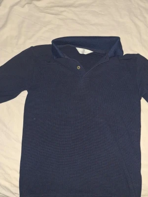 Mörkblå henley från H&M - Säljer en mörkblå henley från H&M med lång ärm och en knapp i halsen. Skjortan är gjord i mjuk bomull och har en klassisk krage. Perfekt för dig som gillar enkel och stilren design.
