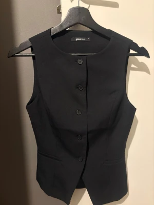 Svart väst från Gina Tricot - Stilren svart väst från Gina Tricot med klassisk knäppning framtill och två fickor. Västen har en figurnära passform och är tillverkad i ett slätt material som ger en snygg look till alla outfits.