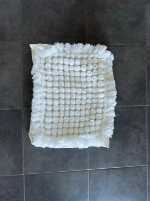 Kuddfodral nya - Säljer en vit rektangulär matta med fluffiga bollar och fransar runt kanten. Mattan har en mjuk och mysig känsla och passar perfekt som dekoration eller till att ha vid sängen. Materialet är textil och färgen är helvit.