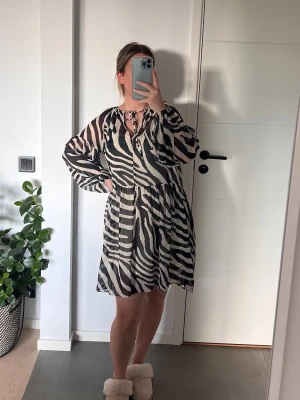 Zebramönstrad kort klänning - Säljer en kort klänning med zebramönster i svart och beige. Klänningen har långärmade, transparenta ärmar och en v-ringad hals med knytband. Perfekt för dig som vill sticka ut med ett djurmönster!