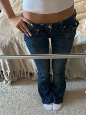 Mörkblå low rise jeans True Religion - Säljer ett par mörkblå low rise jeans från True Religion med utsvängda ben och dekorativa kontrastsömmar. Jeansen har guldiga knappar och detaljer samt klassiska fickor med True Religion-logga bak. 
