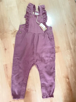 Lila jumpsuit med volangdetaljer, 86, Newbie - Supersöt lila jumpsuit i mjukt, luftigt tyg med volangprydda axelband och knappar i sidorna. Resår vid benslut och en liten ficka bak ger extra stilpoäng. Perfekt för dig som vill ha något bekvämt och trendigt!