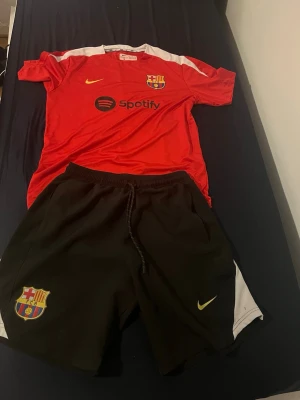 FC Barcelona Nike fotbollskit röd/svart - Säljer ett FC Barcelona fotbollskit från Nike med röd tröja och svarta shorts. Tröjan har klubbmärke, Spotify-logga och vita detaljer på axlarna. Shortsen har klubbmärke och vita paneler på sidorna. Perfekt för träning eller match.