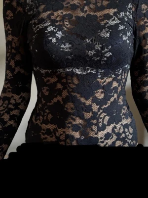 Spets body  - Snygg svart spetsbody med långärmad design och blommigt spetsmönster. Klänningen har en djup, öppen rygg och figurnära passform som framhäver siluetten. Perfekt för dig som vill sticka ut med en elegant och trendig look.