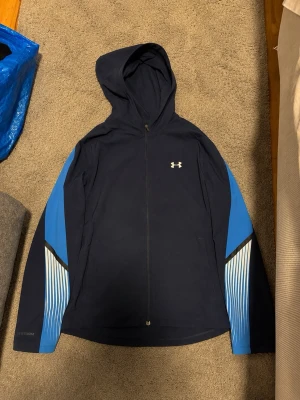 Under Armour Jacka - Snygg marinblå vindjacka från Under Armour med huva och dragkedja. Jackan har blå och vita detaljer längs ärmarna samt logga på bröstet. Perfekt för träning eller när du vill ha en sportig look. Lätt material som andas. Denna är slut såld överallt!