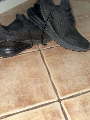 Svarta Nike Air Max 270 sneakers - Säljer ett par helsvarta Nike Air Max 270 sneakers med mesh-överdel och stor synlig Air-enhet i hälen. Skorna har en sportig look med diskret Nike-logga på sidan och snörning framtill. Perfekta för dig som gillar streetwear och bekväma skor.