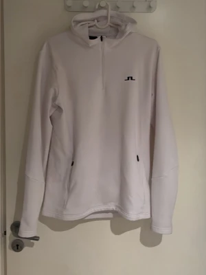 Vit hoodie med halv dragkedja - Säljer en stilren vit hoodie ifrån J.Lindeberg med halv zip framtill och svart logga på bröstet. Två fickor på sidan. Nypris ca 1500kr, knappt använd. Inga skador/fläckar. 