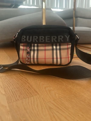 Burberry axelväska med rutigt mönster - Snygg axelväska från Burberry med klassiskt beige, svart och rött rutmönster på framsidan. Väskan har svart kant och justerbar axelrem. Stor logga på ovansidan och dragkedja med rödvit detalj. Perfekt för dig som gillar streetstyle och designeraccessoarer. Helt ny 