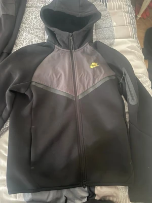 Svart Nike set med huva och dragkedja - Säljer ett svart Nike set med matchande hoodie och byxor. Hoodien har dragkedja, huva och gul Nike-logga på bröstet. Byxorna har resår i midjan och samma logga på benet. Setet är gjort i mjukt och stretchigt material, perfekt för en sportig look.
