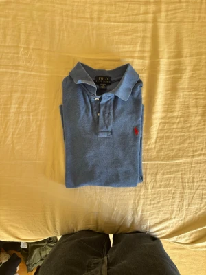 Blå pikétröja från Polo Ralph Lauren - Säljer en klassisk blå pikétröja från Polo Ralph Lauren med korta ärmar och krage. Tröjan har knappar framtill och den ikoniska röda logotypen broderad på bröstet. Perfekt till jeans eller shorts för en avslappnad stil.