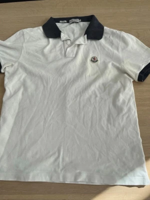 Vit Moncler piké med mörkblå krage - Säljer en klassisk vit pikétröja från Moncler med mörkblå krage och ärmslut. Tröjan har knappslå framtill och Moncler-logga broderad på bröstet. Slim fit-modell som passar perfekt till jeans eller shorts. Sitter bättre som en S/M 