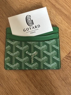 Grön kortfodral  - Snyggt grönt kortfodral med det klassiska mönstret i vitt och grått. Fodralet har flera fack för kort och är tillverkat i tåligt canvasmaterial med läderdetaljer. Perfekt för dig som vill ha en stilren och exklusiv accessoar.