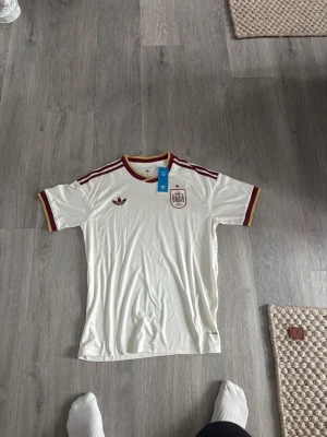 Vit Spanien fotbollströja Adidas - Säljer en vit fotbollströja från Adidas med Spaniens landslagsmärke på bröstet. Tröjan har vinröda och gula detaljer vid krage och ärmslut samt tre vinröda ränder på axlarna. Perfekt för dig som älskar fotboll och vill ha en snygg supportertröja.