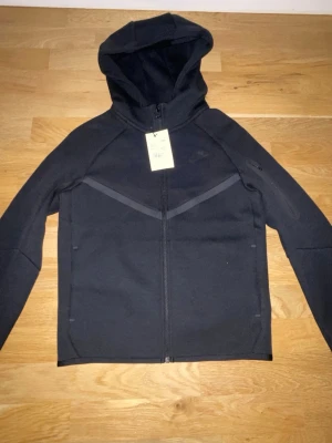 Svart hoodie med dragkedja och luva - Snygg svart hoodie med hel dragkedja framtill och luva. Hoodien har diskreta mörka detaljer i form av ett V-mönster över bröstet. Tillverkad i ett mjukt och bekvämt material, perfekt för en avslappnad stil.
