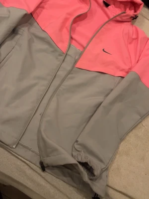 Nike Windbreaker Jacket - This is a Nike windbreaker jacket featuring a two-tone color scheme with a pink upper section and a light gray lower section and sleeves. The jacket has a full front zipper, a hood with drawstrings, and elastic cuffs. The Nike logo is visible on the chest.