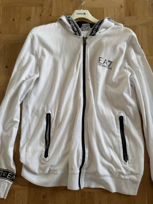 Vit hoodie från EA7 Emporio Armani - Snygg vit hoodie från EA7 Emporio Armani med dragkedja framtill och svarta detaljer. Logga på bröstet och tryck på huvan. Två fickor med dragkedja och ribbade muddar. Perfekt för en clean och sportig look.