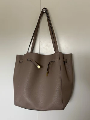 Beige shoppingväska i skinnimitation - Stilren shoppingväska i beige skinnimitation med två långa axelband och guldfärgade detaljer. Väskan har en enkel, rymlig design och stängs med ett dekorativt band framtill. Perfekt för dig som vill ha plats för allt du behöver under dagen.