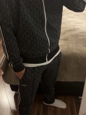 Lacoste Mörkgrått mönstrat tracksuit-set - Säljer ett mörkgrått tracksuit-set med jacka och byxor. Setet har ett rutigt mönster, vita ränder längs ärmar och ben samt vita dragkedjor och snören. Både jackan och byxorna har en liten grön logga på bröstet och höften.