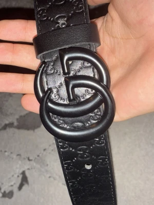 Svart bälte  - Snyggt svart bälte i konstläder svart mönster och metallspänne. Bältet har flera hål för justering och en stilren design som passar till många olika outfits. 116cm