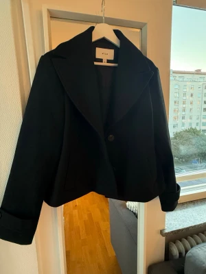 Svart kort kappa från Vila - Stilren svart kappa från Vila med breda slag och två knappar framtill. Modellen är kort och har uppvikta ärmslut.