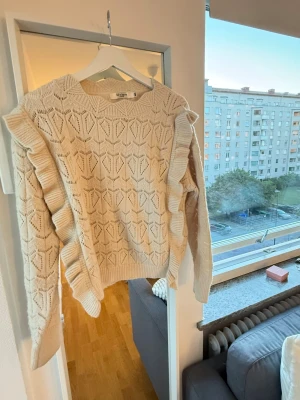 Beige stickad tröja med volang - Säljer en beige stickad tröja med dekorativa volanger längs ärmarna och ett snyggt hålmönster över hela plagget. Tröjan har rund halsringning och ribbade muddar vid ärmslut och nederkant. 