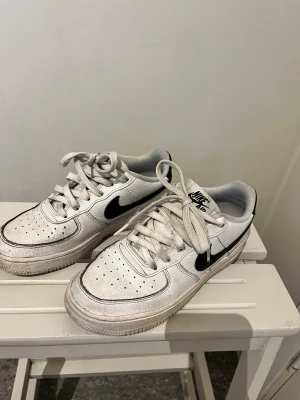 Nike Air Force 1 vita sneakers - Klassiska vita Nike Air Force 1 sneakers med svart swoosh på sidan. Skorna har perforerad tå, vita skosnören och en robust sula. Tillverkade i läder med textildetaljer och låg profil. Perfekta för en streetwear-look.