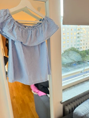 Blåvit offshoulder randig topp - Säljer en söt offshoulder topp med volangkant. Toppen är blåvit med smala ränder och har en luftig passform. Perfekt för varma dagar och ger en fräsch sommarkänsla. Tillverkad i mjuk bomull som känns skön mot huden.