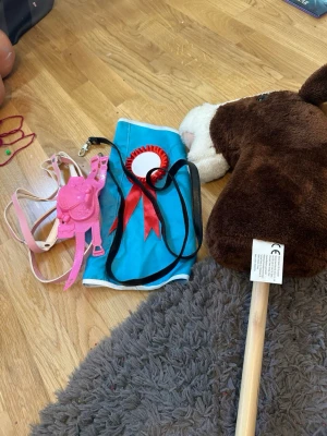 Käpphäst med tillbehör - Säljer två käpphästar, en brun med svart man och en mörkbrun med vit mule. Tillbehör som sadel i rosa i plast,  blått täcke med rosett och svart tygtygel ingår. Perfekt för lekar och tävlingar hemma eller utomhus.