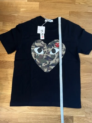 Svart T-shirt comme des garcons cammoflaugehjärta - Snygg svart t-shirt från Comme des Garçons PLAY med stort hjärta i camouflage-mönster och ikoniska ögon på bröstet. Liten röd hjärtdetalj i hörnet. Klassisk passform och rund hals. Perfekt för dig som gillar streetwear och statement-plagg.