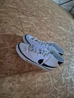 Vita höga sneakers med svart swoosh - Snygga vita sneakers med högt skaft och svart swoosh på sidan. Skorna har vita snören, svart rand längs sulan och svart detalj vid hälen. Tillverkade i canvasmaterial för en klassisk look.