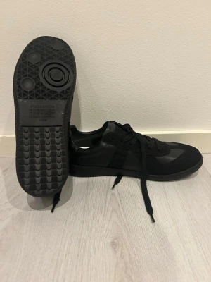 Svarta sneakers Maison Margiela - Snygga helsvarta sneakers från Maison Margiela med klassisk låg siluett. Skorna har en mix av mocka och läderdetaljer, svarta skosnören och gummisula med unikt mönster undertill. Perfekta för dig som gillar minimalistisk och stilren design. OBS! De är helt nya o aldrig använda. skriv om ni har frågor!