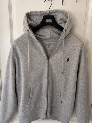 Grå zip hoodie från Polo Ralph Lauren - Snygg grå zip hoodie från Polo Ralph Lauren med klassisk huva och dragsko. Broderad marinblå logga på bröstet och två fickor framtill. Tillverkad i mjuk bomullsblandning, perfekt för en chill och avslappnad stil. Går ej under 275