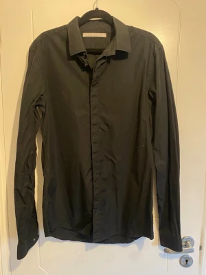 Svart skjorta från Tiger of Sweden - Klassisk svart skjorta från Tiger of Sweden med lång ärm och knappar framtill. Skjortan har en stilren design och är tillverkad i ett mjukt bomullstyg som passar perfekt till olika outfits.