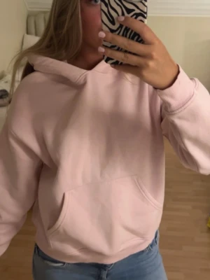 Ljusrosa hoodie med ficka - Säljer en ljusrosa hoodie med huva och stor magficka. Hoodien har en avslappnad passform och ribbade muddar vid ärmar och nederkant. Perfekt för en chill och bekväm stil.