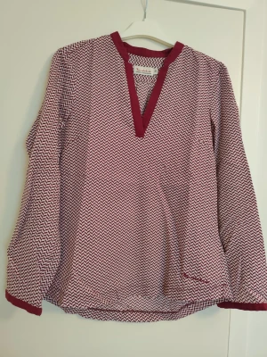 Mönstrad blus med vinröda detaljer - Säljer en snygg blus med vinrött och vitt zigzag-mönster. Blusen har lång ärm, v-ringad hals med vinröda kanter och slitsar vid ärmslut och nederkant. Materialet känns lätt och luftigt, perfekt för en trendig look.