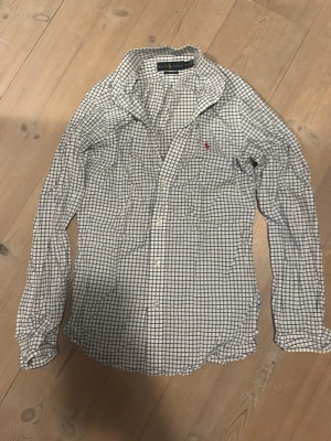 Rutig skjorta från Ralph Lauren - Klassisk vit skjorta från Ralph Lauren med blått rutmönster och röd broderad logga på bröstet. Slim fit-modell med långa ärmar och knappar framtill. Tillverkad i mjuk bomull som känns skön mot huden. Perfekt för en clean och stilren look.