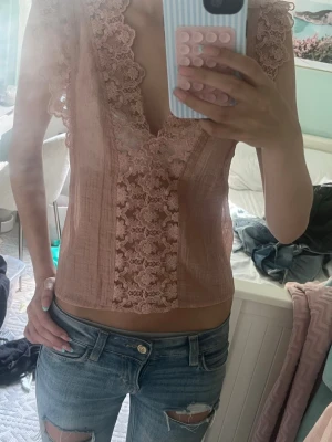 Rosa spetsblus med djup v-ringning - Söt ljusrosa blus med djup v-ringning och breda spetsdetaljer längs halsringningen och framtill. Ärmlös modell med romantisk känsla och lätt transparent tyg. Perfekt att styla med jeans eller kjol för en trendig look.