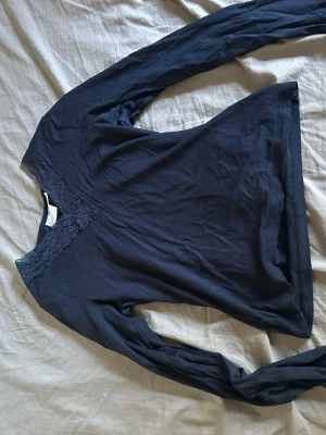 Mörkblå långärmad topp med spets - Mörkblå långärmad topp med v-ringning och dekorativ spetsdetalj vid halsen. Tunn och mjuk i materialet, perfekt för lager på lager eller som den är. Enkel men ändå elegant design som passar till mycket.