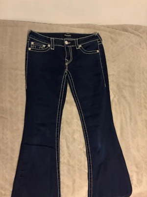 Mörkblå bootcut jeans True Religion - Säljer ett par mörkblå bootcut jeans från True Religion med kontrasterande vita sömmar och klassisk femficksdesign. Jeansen har låg midja och utsvängda ben, vilket ger en retro och trendig look. Perfekta för dig som gillar statement-jeans!