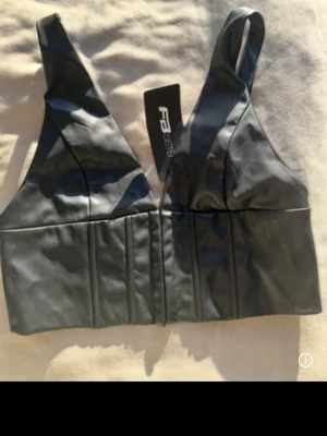 Svart faux skinn korsettopp - Snygg svart korsettopp i syntetiskt skinn med breda axelband och markerade sömmar framtill. Toppen har en croppad passform och djup v-ringning, perfekt för att lyfta vilken outfit som helst. Helt ny med prislapp kvar! 🌷Spana även in mina andra plagg!🤗