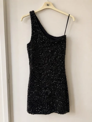 Svart glittrig oneshoulder klänning - Snygg svart kortklänning med glittriga paljetter och oneshoulder-design. Klänningen har en smal axelrem på ena sidan och är ärmlös på andra. Perfekt för dig som vill sticka ut med en trendig och festlig look.
