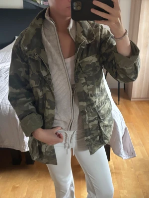 Militär färgad jacka  - Snygg kamouflagemönster. Jackan har dragkedja och knappar framtill, två stora fickor och justerbar midja
