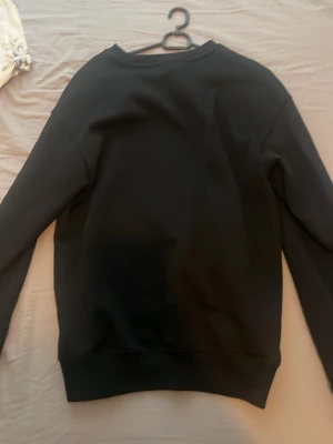 Svart basic hoodie utan huva - En enkel svart hoodie i mjukt material med rund hals och långa ärmar. Modellen är rak och har ribbade muddar vid ärmslut och nederkant. Perfekt för en clean och avslappnad stil.